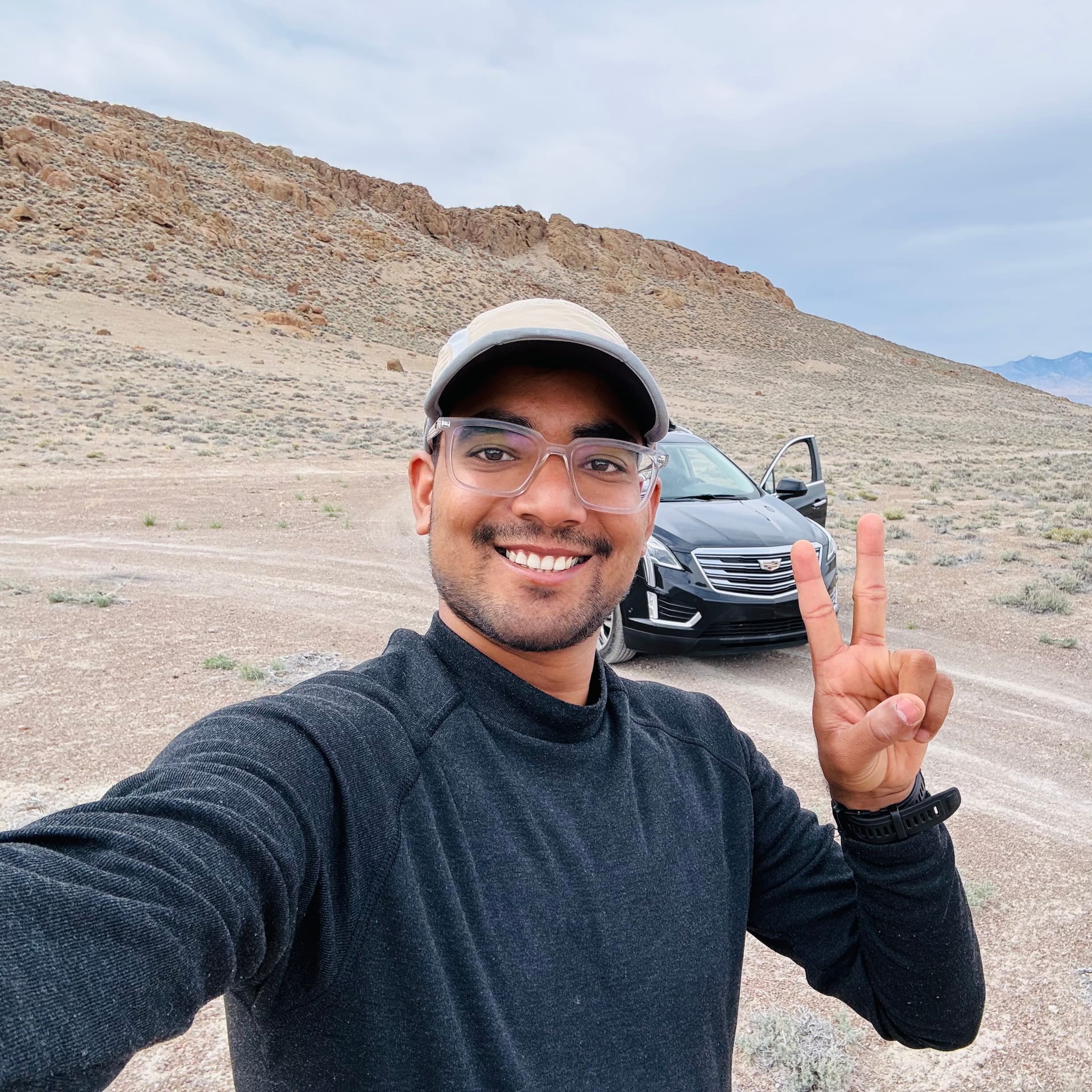 Selfie at the Volcano Peak trailhead with Cadillac parked on a desert dirt road, rocky hills behind